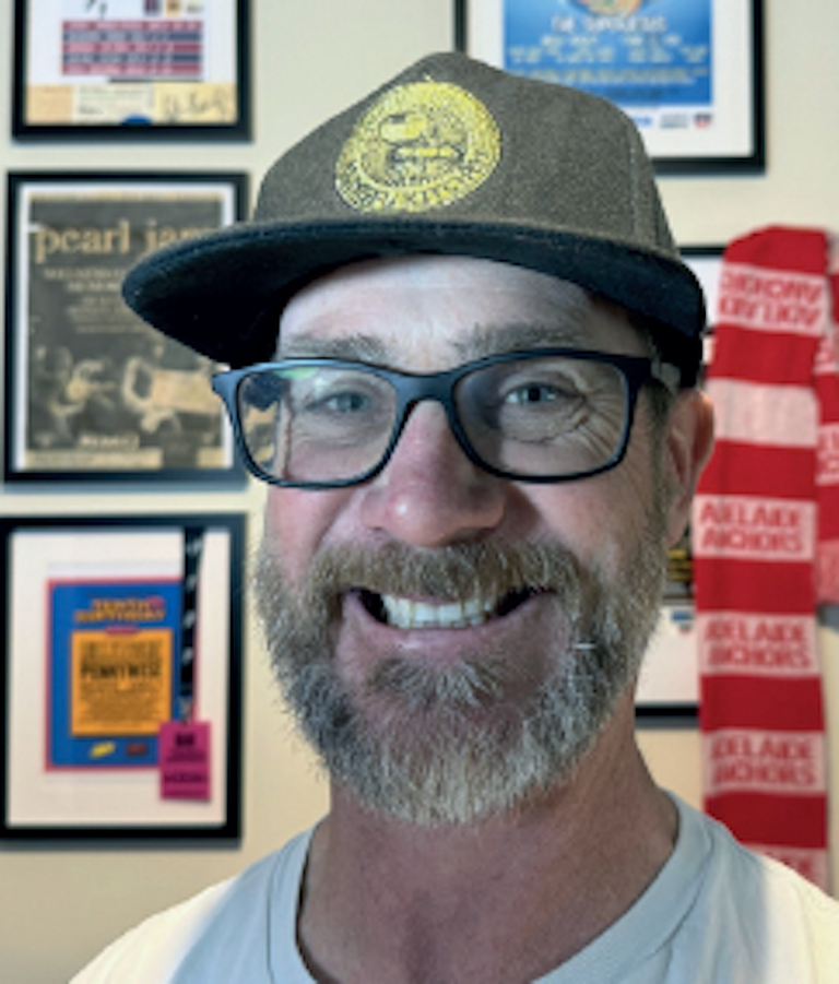 Portrait of Shannon Mowling wearing glasses and a cap, smiling in front of framed music memorabilia and a red Adelaide Anchors scarf.